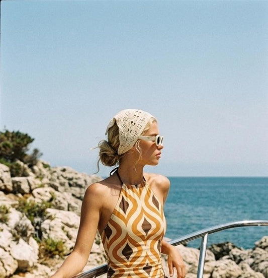 May include: A cream-coloured crochet headscarf with a delicate openwork pattern and a halter-neck top featuring a repeating brown, tan, and white geometric wave print. The outfit is paired with thick-rimmed white sunglasses.